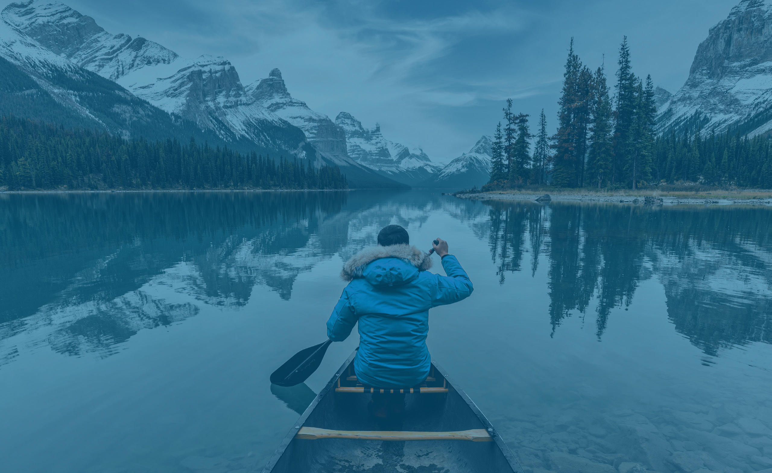 A person in a canoe on a still lake looking at a forest and snow-capped mountains, viewed from behind.