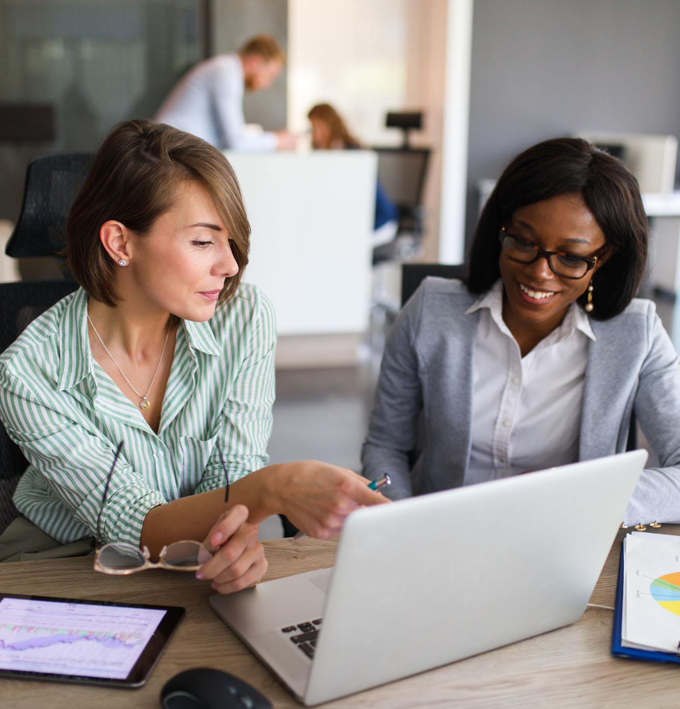 Une femme d’affaires noire et une femme d’affaires blanche travaillent avec une tablette électronique et regardent l’écran d’un ordinateur portable.
